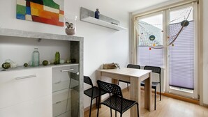 Un comedor con una mesa y sillas, una estantería con objetos decorativos, una ventana con persianas y una hamaca.