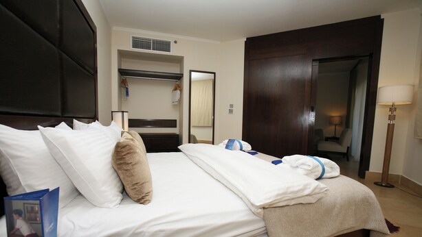 A hotel room with a large bed, a wardrobe, a mirror, a bedside table with a lamp, and a small table with a brochure. - Grand Tala Bay Resort (Aqaba)