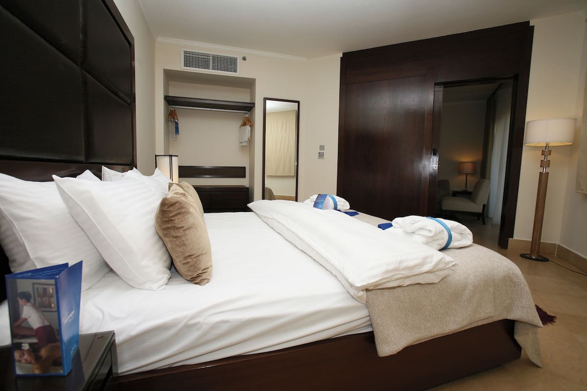 Habitación de hotel con una cama grande, un armario, un espejo, una mesita de noche con una lámpara y una pequeña mesa con un folleto.