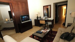 A hotel room with a TV, desk, chair, sofa, and a red patterned rug. - Grand Tala Bay Resort (Aqaba)