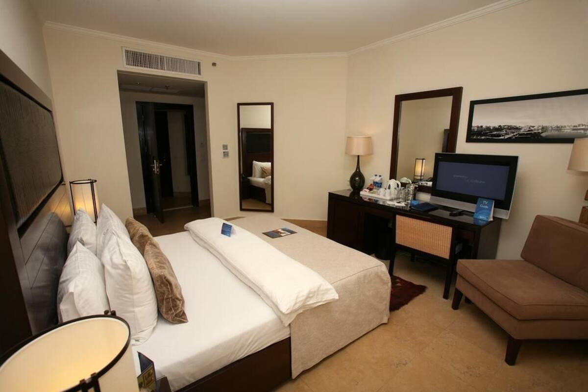 Una habitación de hotel con una cama, un escritorio con televisor, una silla y un espejo.