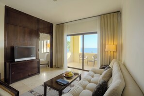 A living room with a flat-screen TV, a sofa, a coffee table with fruits, and a view of the balcony. - Grand Tala Bay Resort (Aqaba)