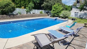 A rectangular swimming pool with a clear blue waterline, surrounded by a white picket fence and lush greenery. There are two lounge chairs with blue and white striped cushions and a metal chair with a grey cushion.