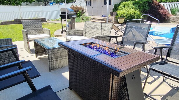 An outdoor patio with wicker furniture, a fire pit, and a pool in the background.