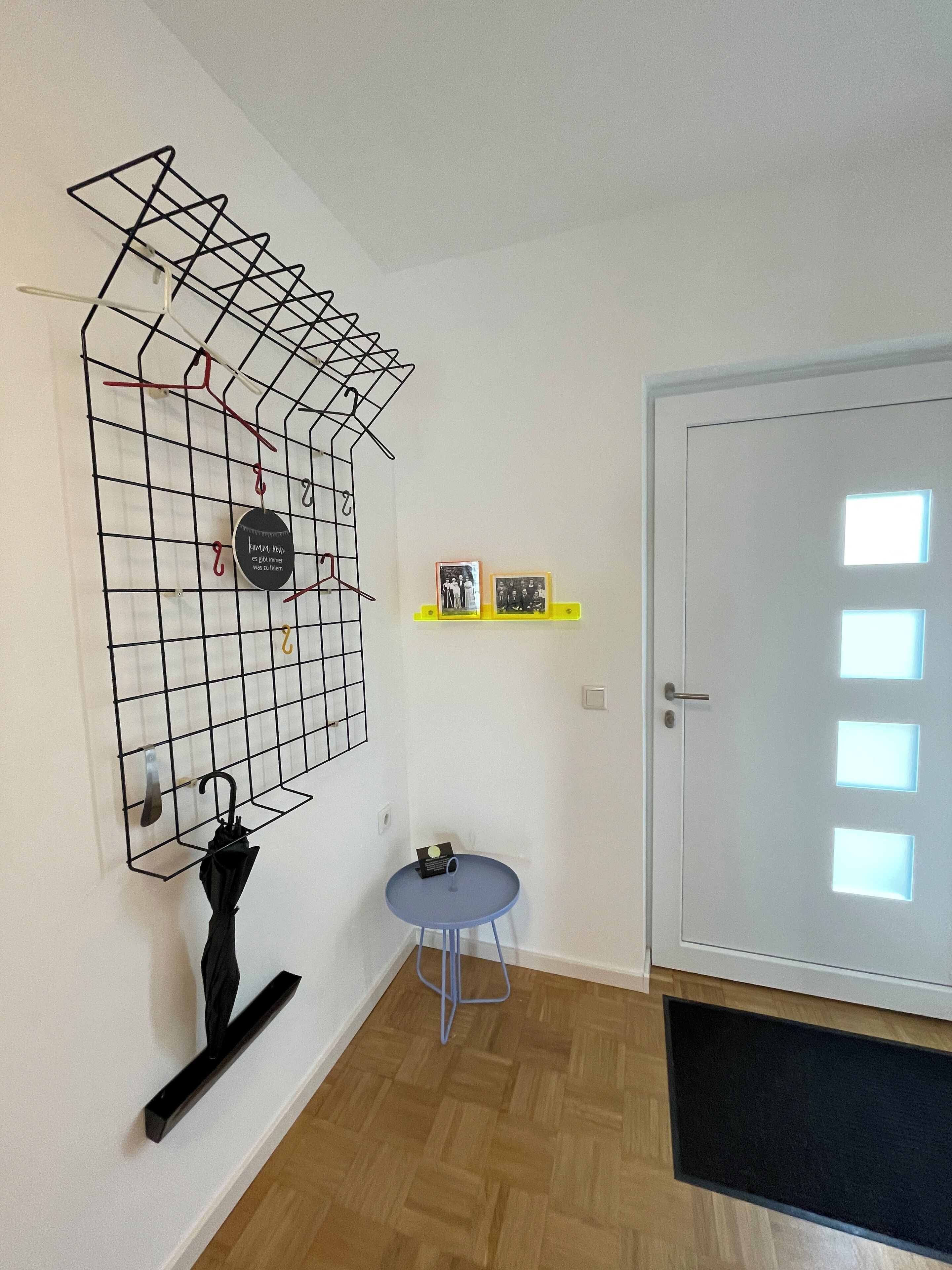 A room with a wooden floor, a white door with a frosted glass panel, a small round table with a blue base, a black metal rack with hangers, and two framed pictures on a yellow shelf.