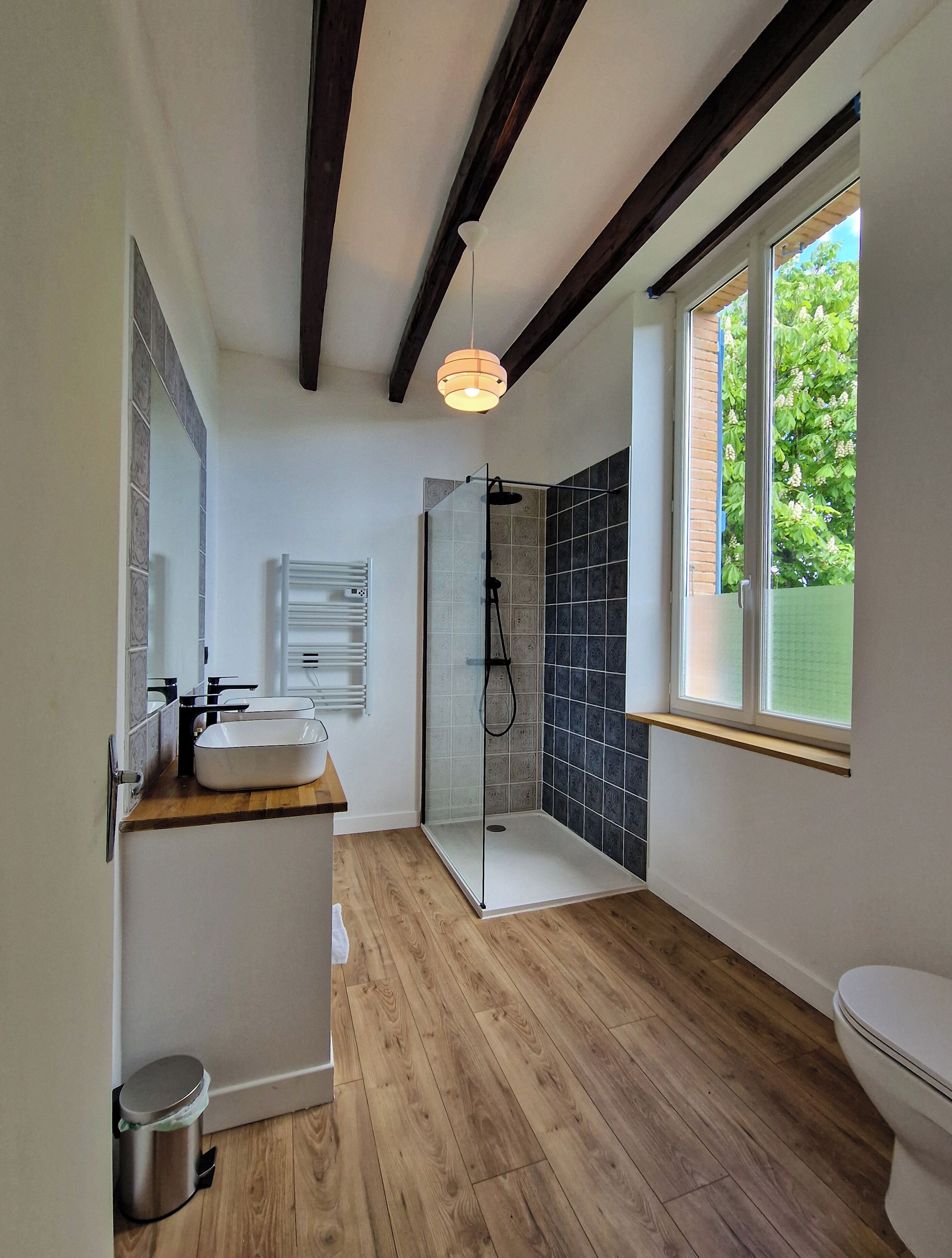 A modern bathroom with a wooden vanity, a glass shower enclosure, and a window providing a view of greenery outside.