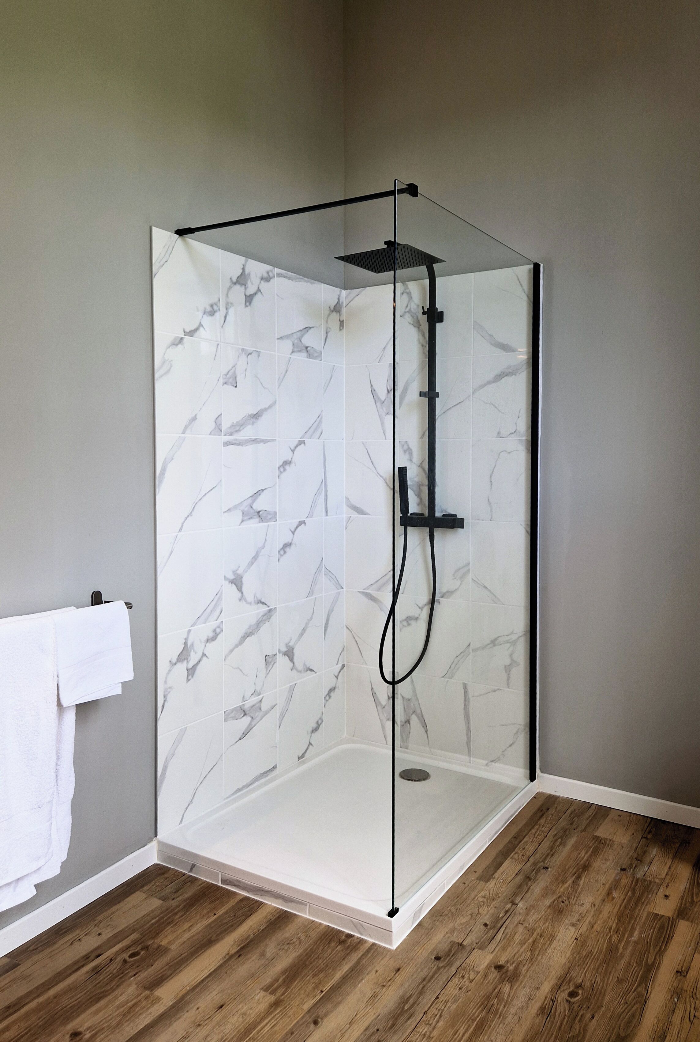 Une douche fermée par des parois de verre, avec un mur au motif de marbre, un plancher en bois et une serviette blanche accrochée au mur.