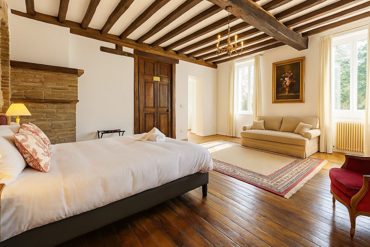 Een slaapkamer met een groot bed, houten balken, een stenen open haard, een bank en een schilderij.