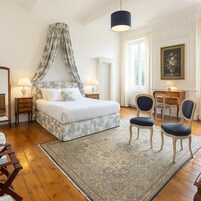 Un dormitorio con cama con dosel, dos mesitas de noche con lámparas, un escritorio con silla y un ventanal con cortinas.