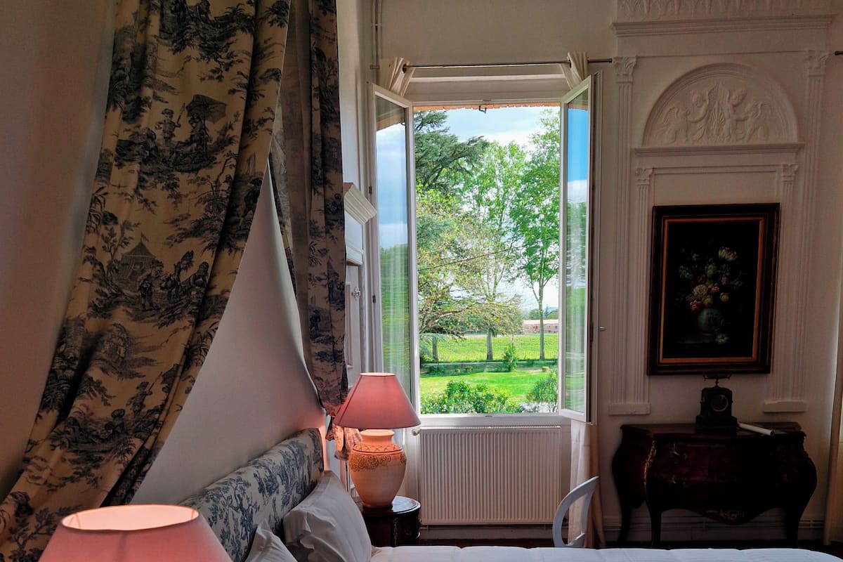 Een slaapkamer met een bed, twee bedlampen, een raam met uitzicht op groen, een radiator en een ingelijst schilderij aan de muur.