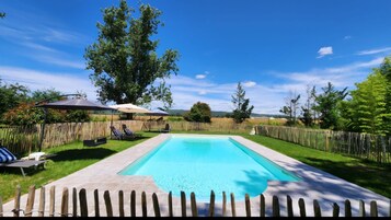 Una piscina rectangular con un deck de madera que la rodea, una valla de madera y dos sillones de jardín con cojines a rayas.