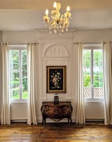 Una habitación con candelabro, chimenea con un cuadro encima y ventanas grandes con cortinas.