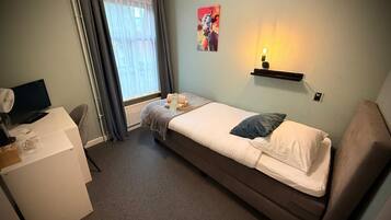 Een kleine eenpersoonskamer met een bureau, een computer, een raam met gordijnen, een wandplank met verlichting, en een schilderij.