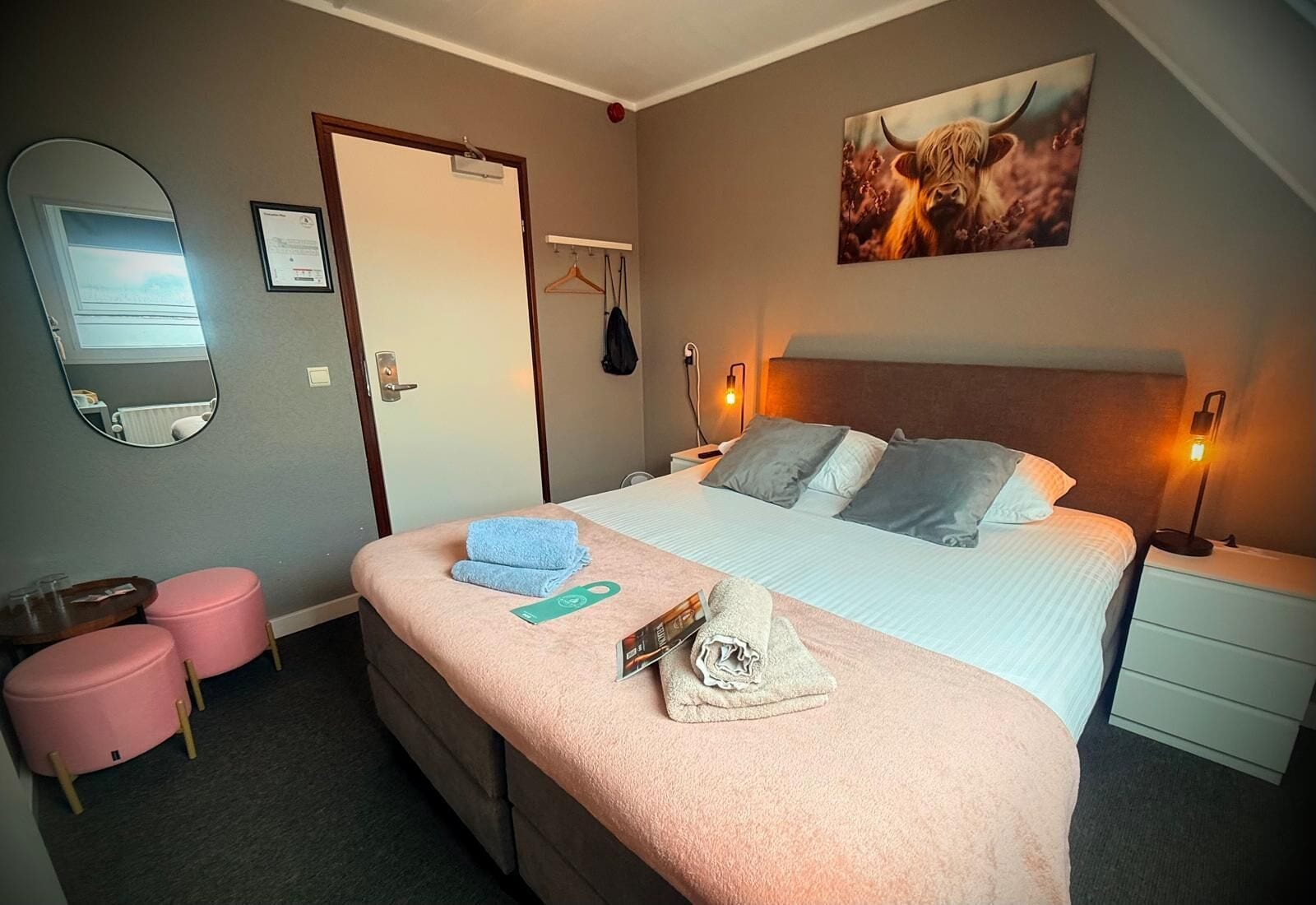 A hotel room with a bed, bedside tables, a mirror, a coat rack, and a painting on the wall.