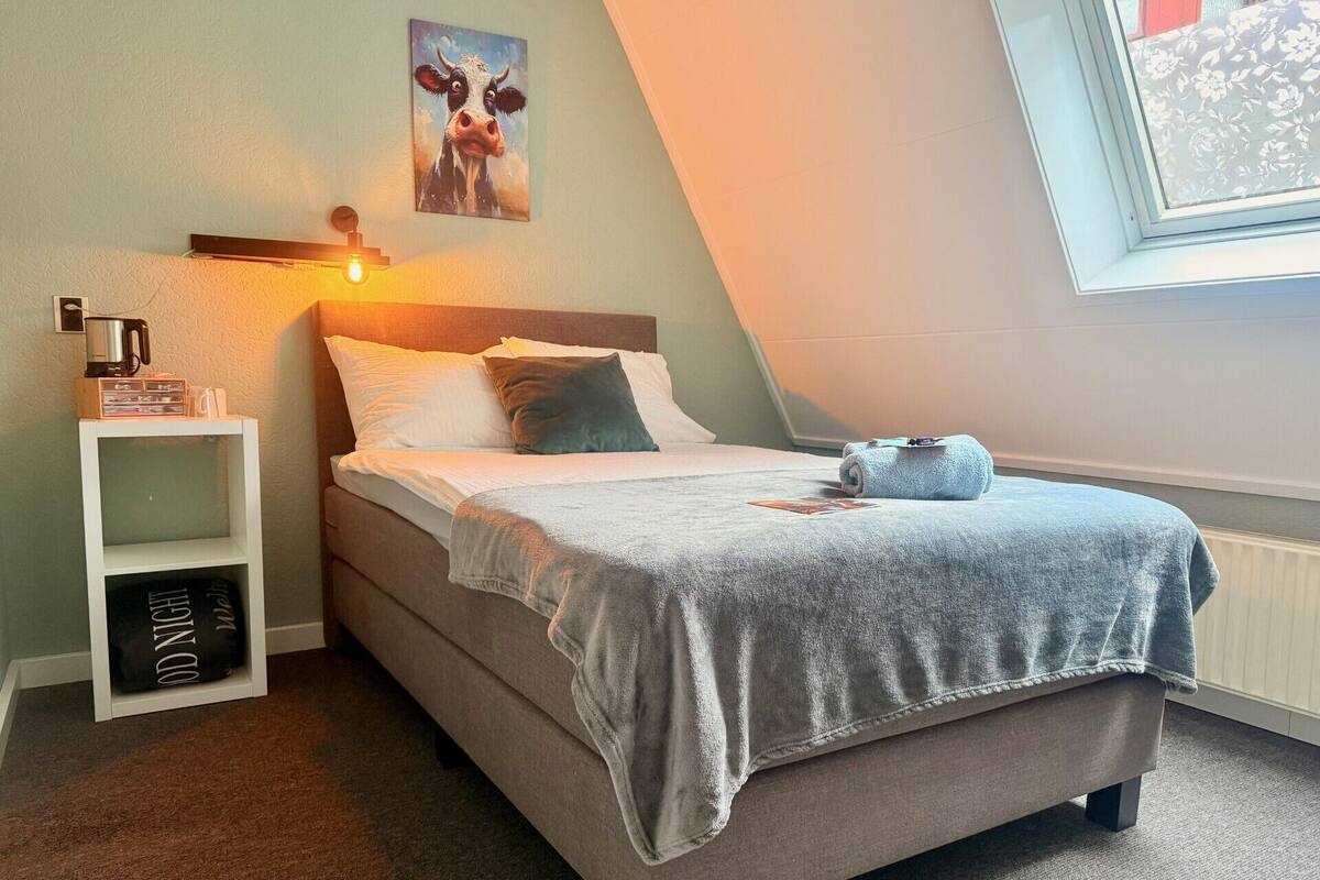 A bedroom with a bed, a nightstand, a window, and a painting on the wall.