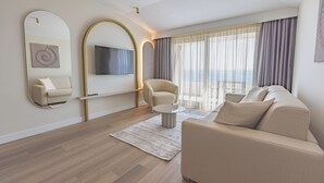 Una sala de estar moderna con sofá, sillón y una mesa de centro redonda. Hay un televisor de pantalla plana montado en la pared y un ventanal con cortinas.