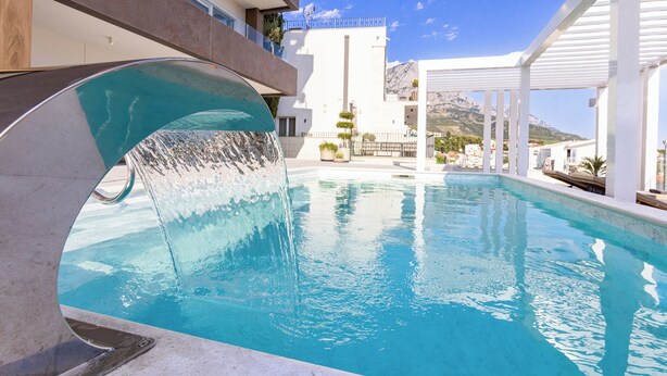 Una piscina moderna con cascada, rodeada de arquitectura blanca y vistas a la montaña.