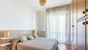 A bedroom with a bed, bedside table, lamp, patterned curtains, and wooden wardrobe.