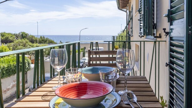 Een houten tafel met kleurrijke borden en wijnglazen, op een balkon met uitzicht op zee.