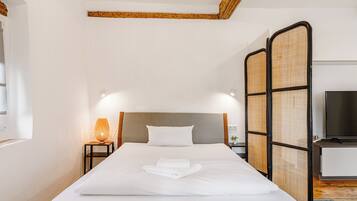 Een net opgemaakt bed met witte lakens en een gevouwen handdoek, een bedlampje, een televisie op een standaard en een geweven scheidingswand.