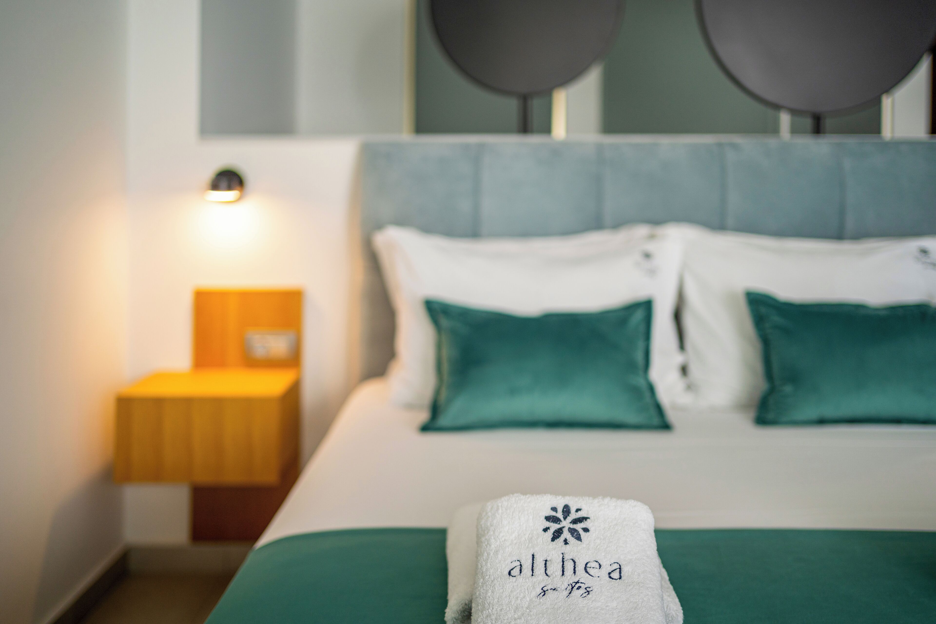 A neatly made bed with a white and green bedspread, two teal pillows, and a white towel with 'althea spa' embroidered on it.