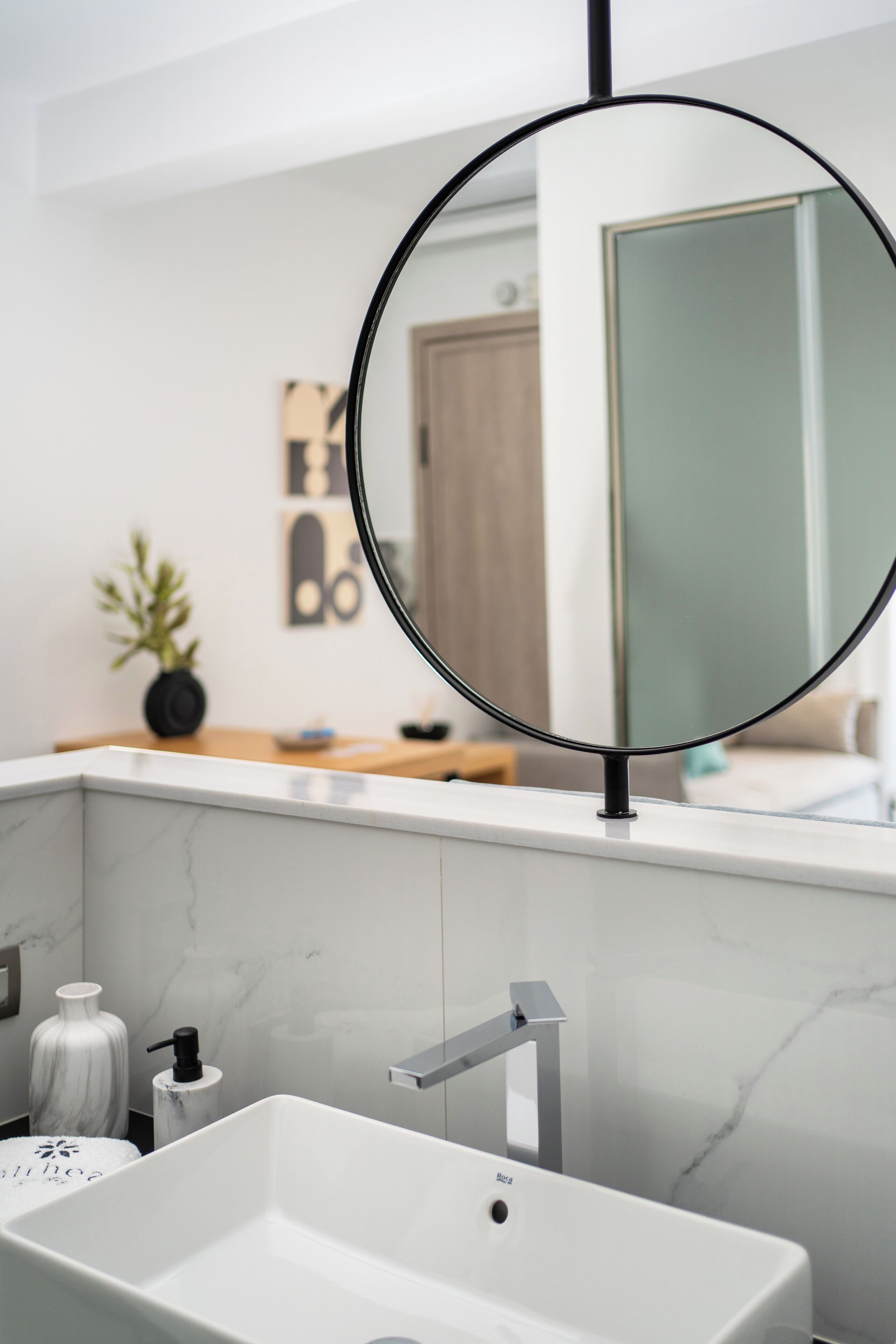 Un baño moderno con un lavamanos blanco, un espejo circular negro y un salpicadero de mármol.