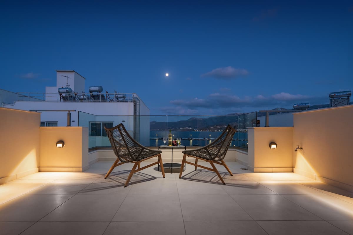 A rooftop terrace with two wicker chairs and a table, overlooking a cityscape and mountains.