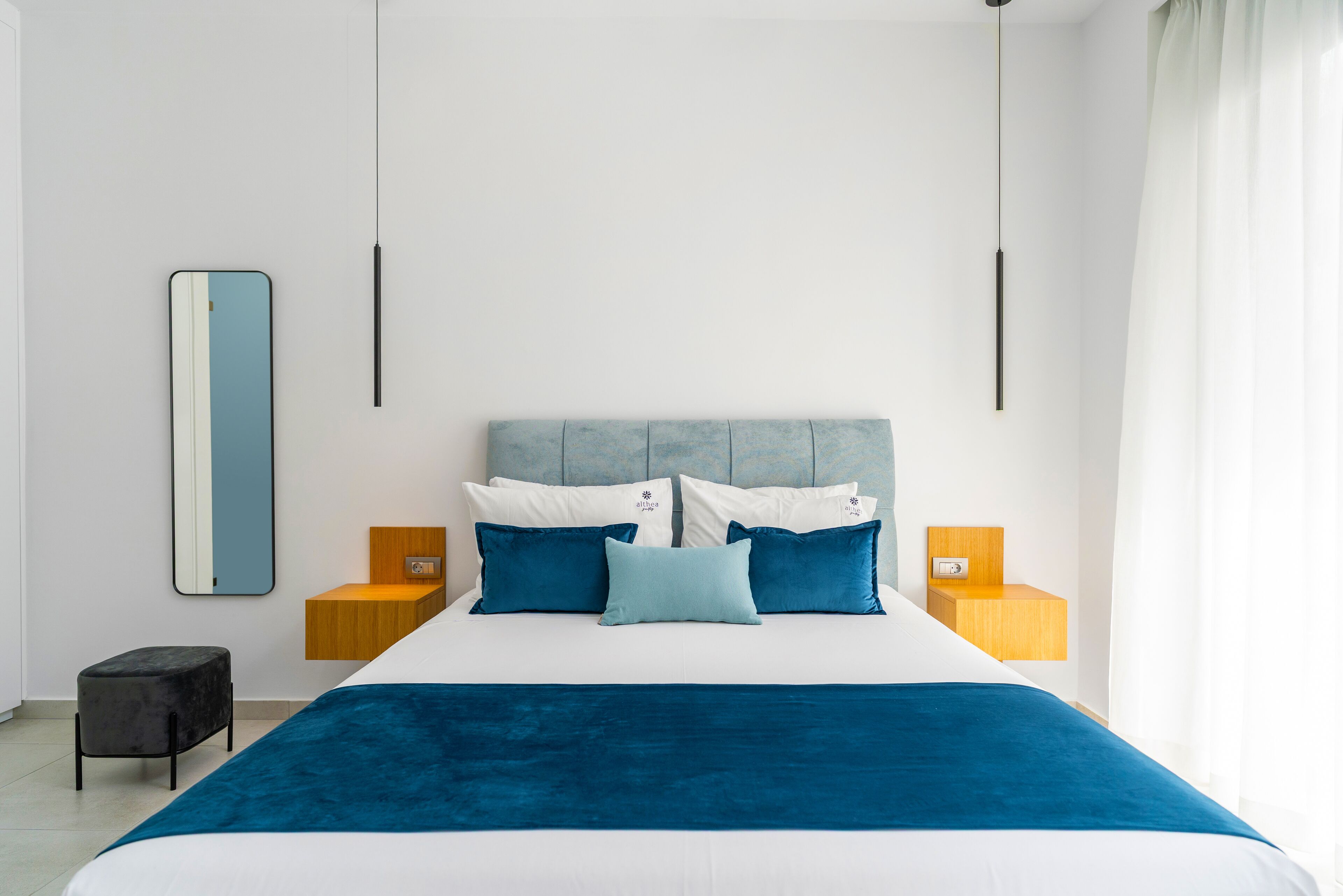 A modern bedroom with a large bed, a headboard, two bedside tables, a nightstand, a mirror, and a small stool.