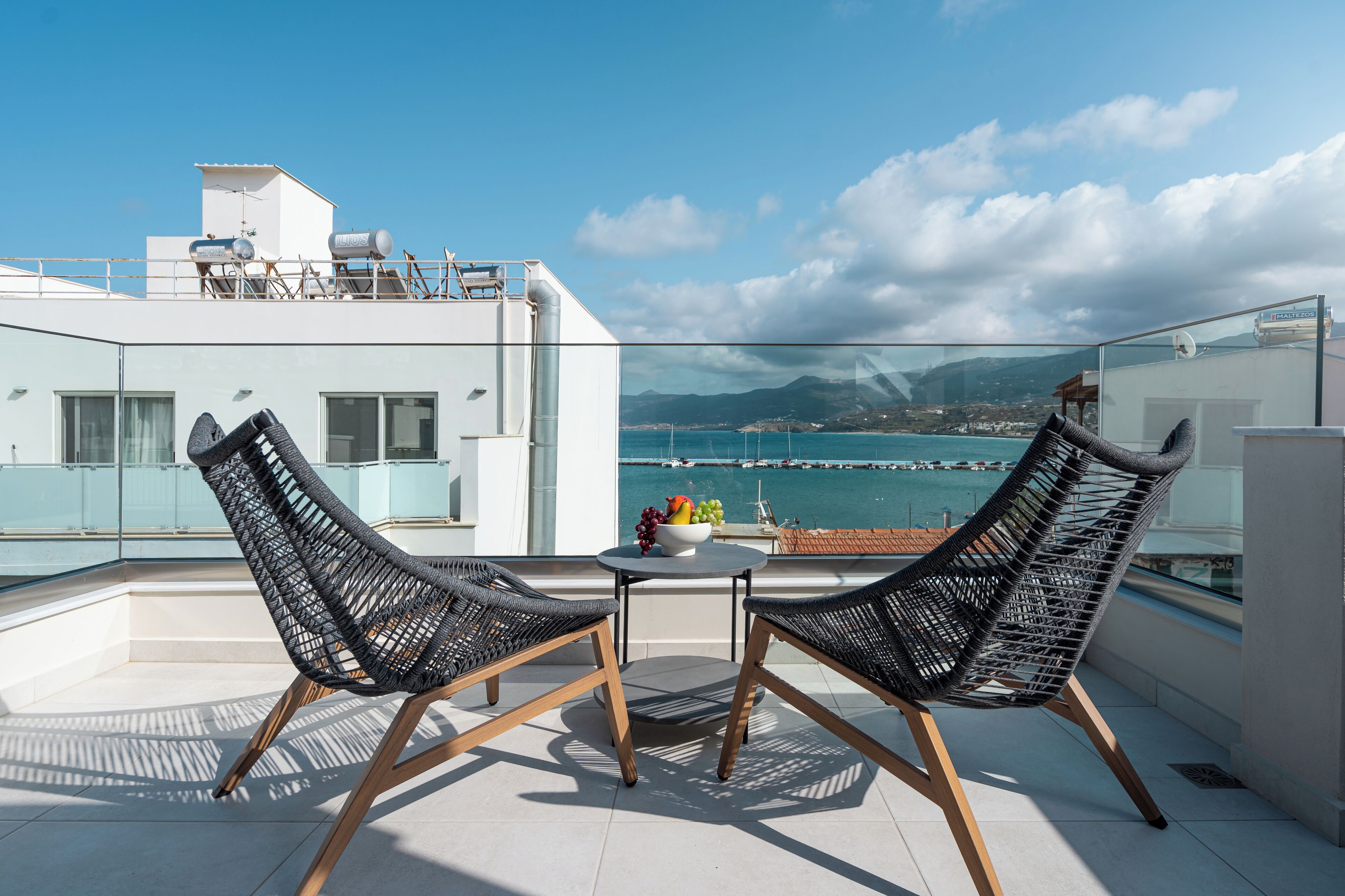 A balcony with two wicker chairs, a small table with fruit, and a view of the sea and mountains.