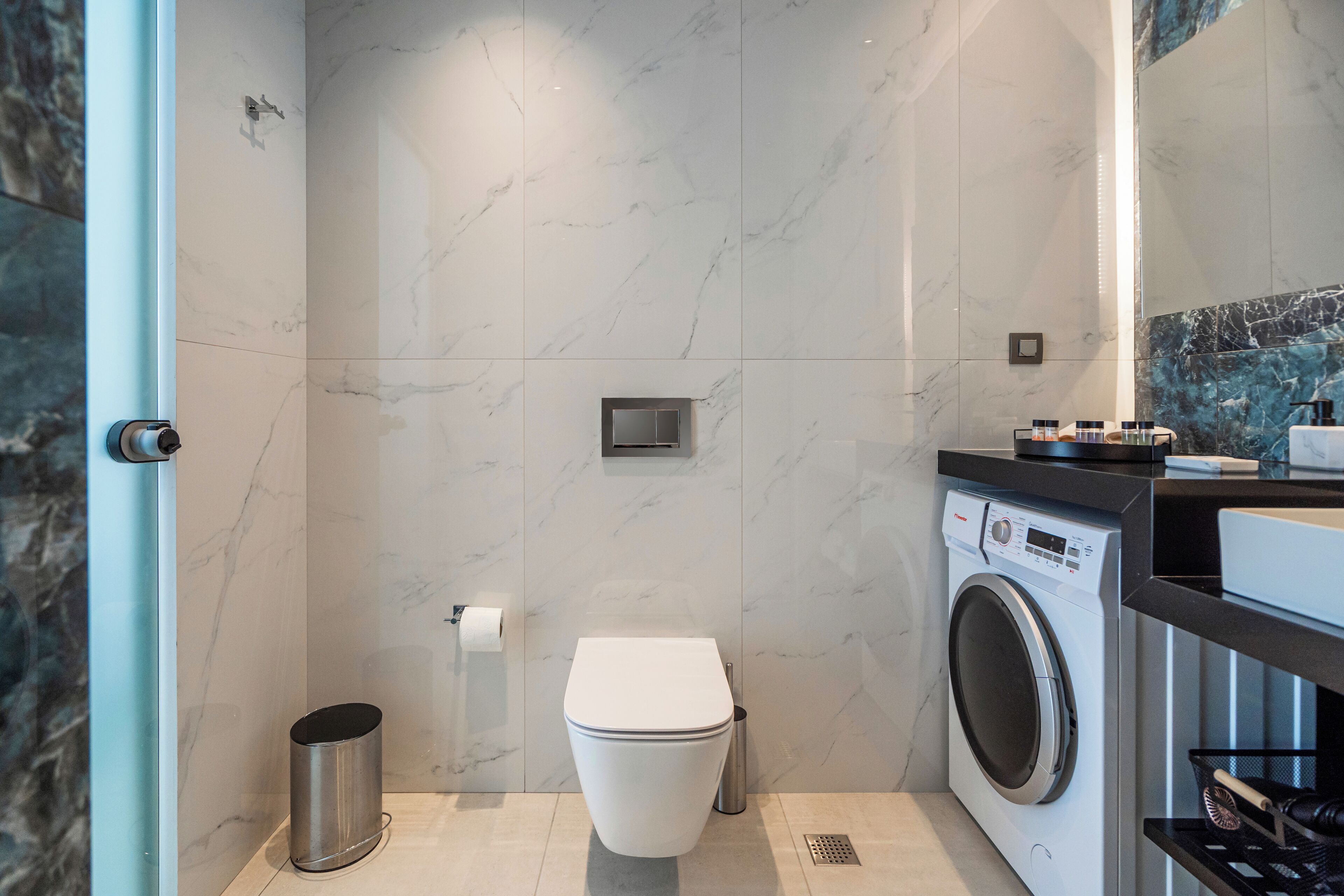 A modern bathroom with a marble wall, a toilet, a washing machine, and a trash can.