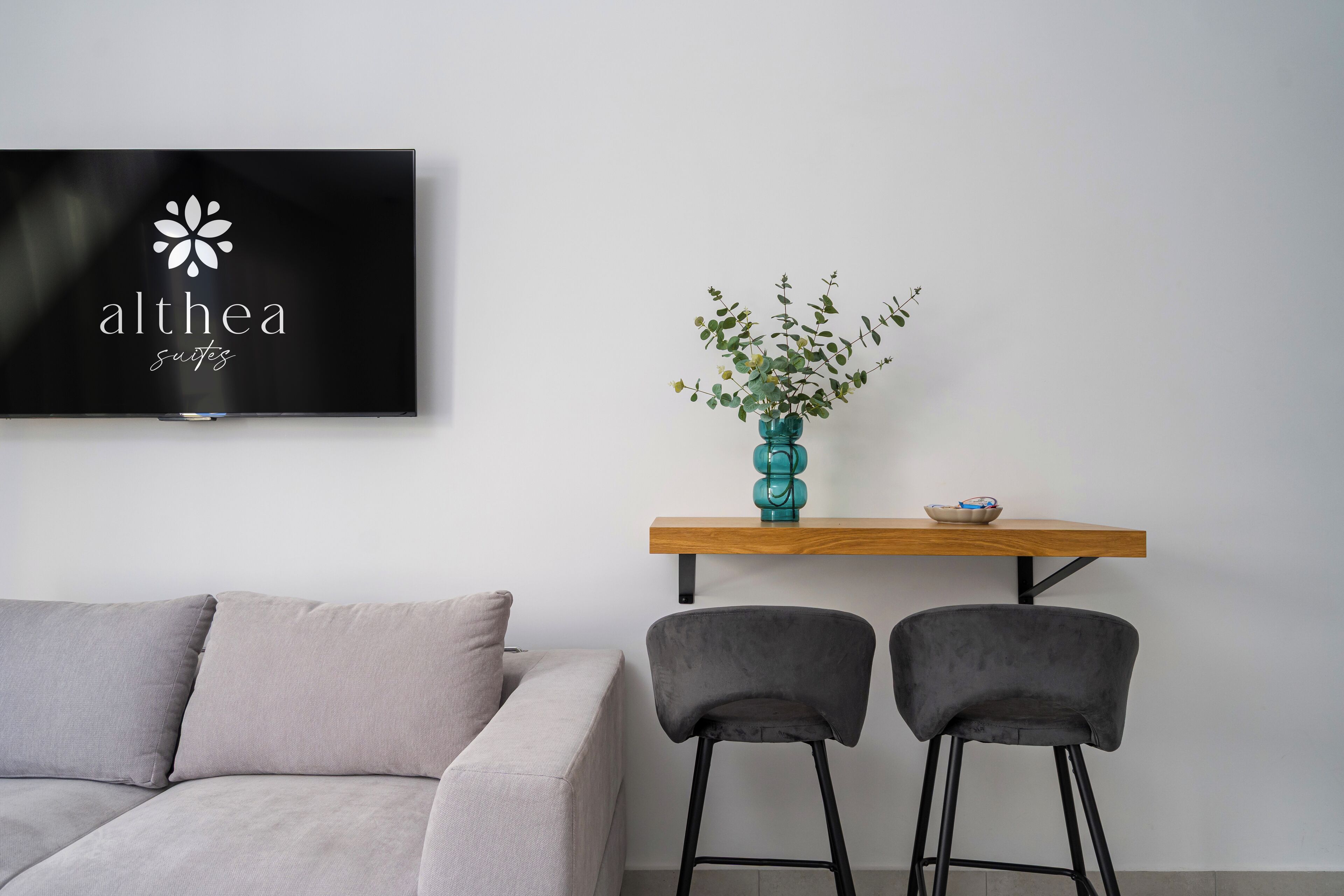 Ett modernt vardagsrum med en grå soffa, två svarta barstolar, ett träbord med en turkos vas och en skål, samt en platt-TV som visar programmet ”Althea Suites”.
