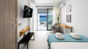 A modern hotel room with a bed, a TV, a desk, a chair, and a view of the sea. - Althea Suites (Sitia)