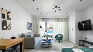 A modern hotel room with a sofa, a dining table, a TV, and a bed with towels. - Althea Suites (Sitia)
