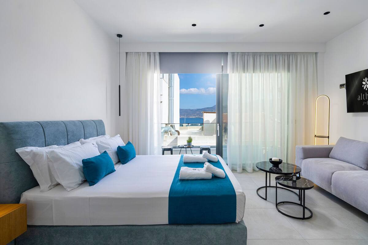 A modern hotel room with a large bed, a sofa, a small table, and a balcony with a view.