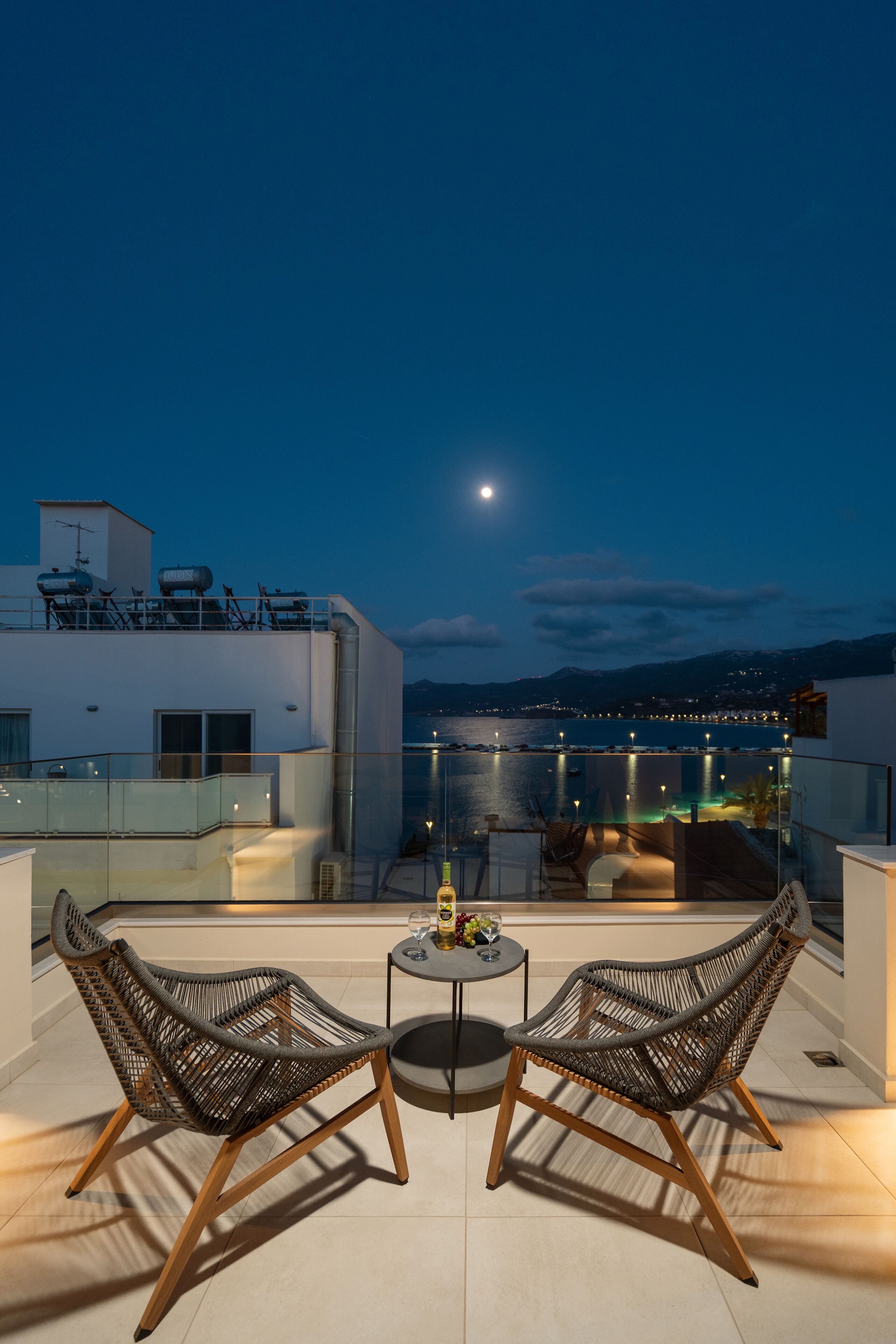 A rooftop terrace with two wicker chairs, a small table, and a view of a body of water and city lights at night.