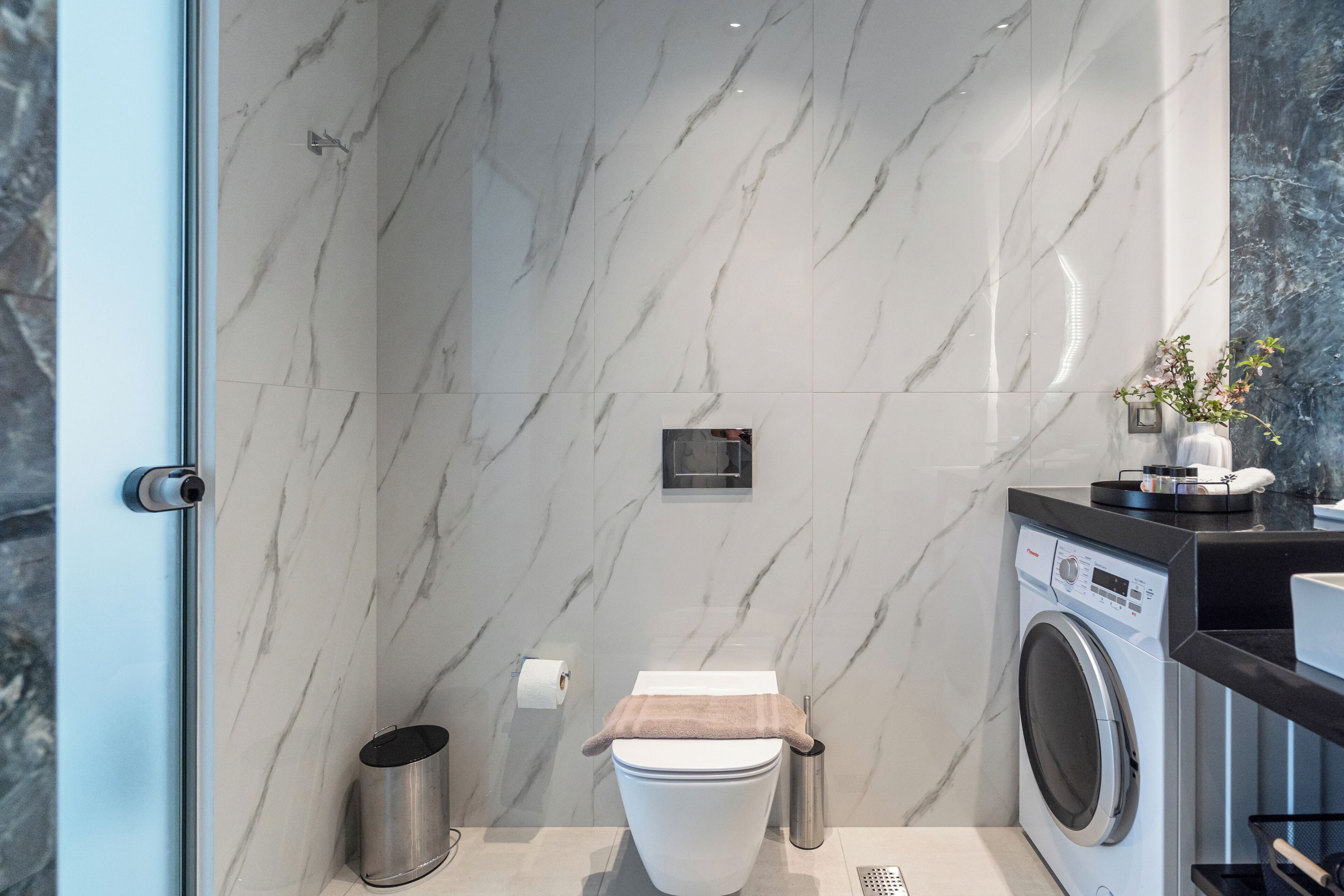 A modern bathroom with a marble wall, a toilet, a washing machine, and a trash bin.