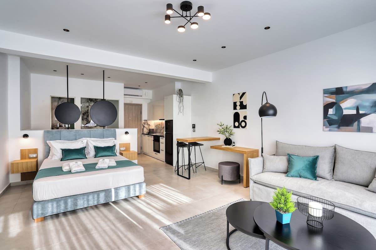 A modern hotel room with a bed, a sofa, a dining area, and a kitchen in the background.