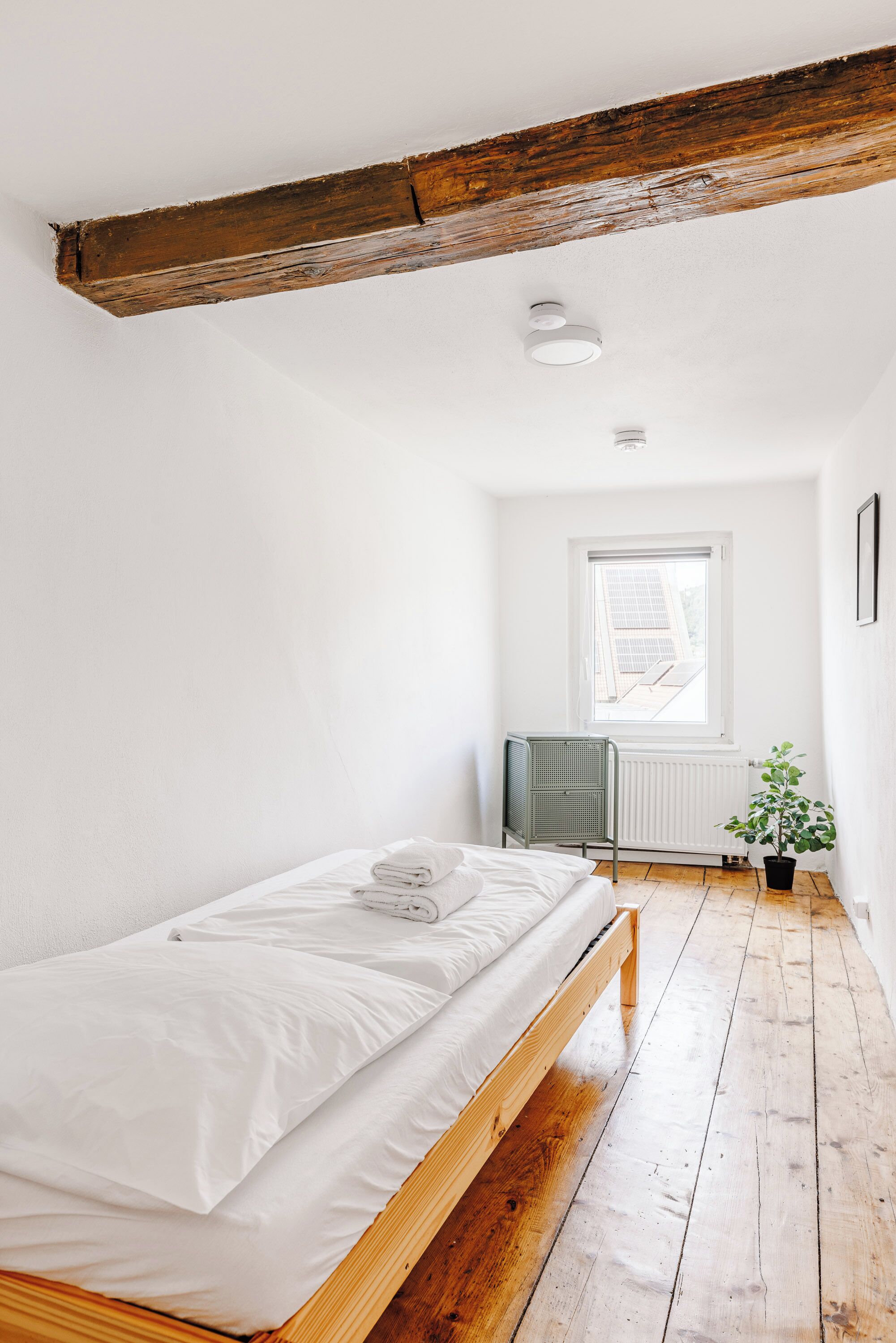 A small, bright room with a wooden floor, a single bed with white linens, a radiator, and a window with a view of the outside.