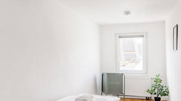 A small, bright room with a wooden floor, a single bed with white linens, a radiator, and a window with a view of the outside.