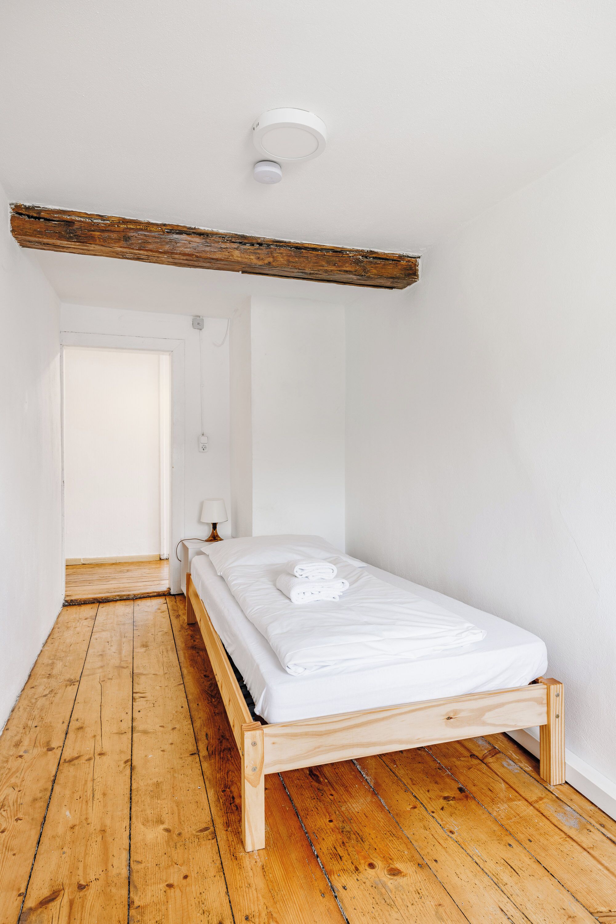A small, bright room with a wooden floor, a simple bed, and a white ceiling.
