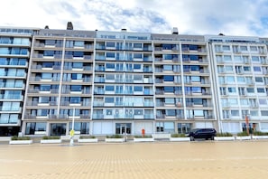 A multi-story residential building with balconies, a gated entrance, and a paved area in front.