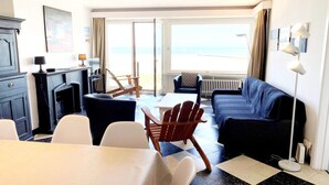 A room with a sofa, chairs, a table, a fireplace, and a view of the beach. - Seaview Apartment in Nieuwpoort (Nieuwpoort)