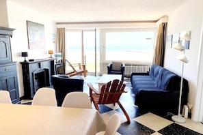 A room with a sofa, chairs, a table, a fireplace, and a view of the beach.