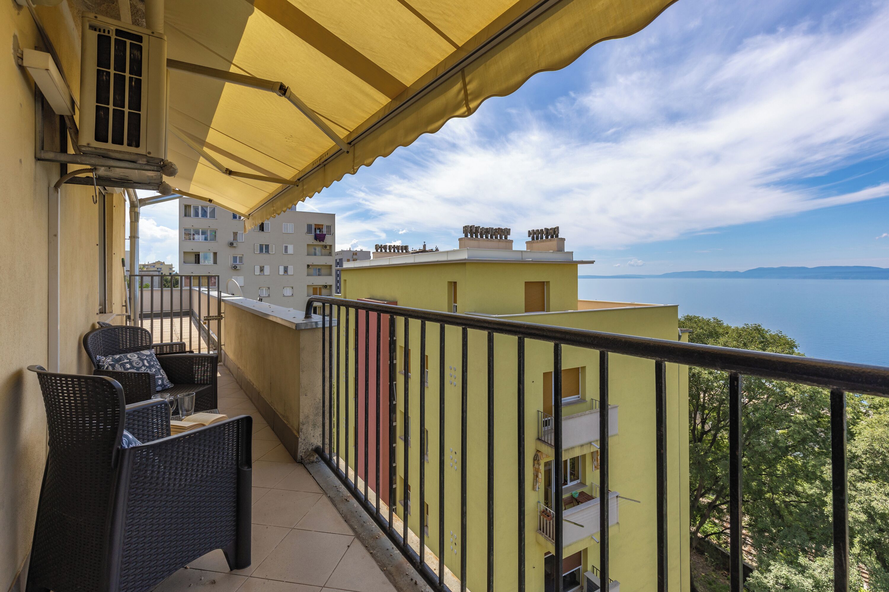 A balcony with wicker furniture, a yellow awning, and a view of buildings and a body of water.