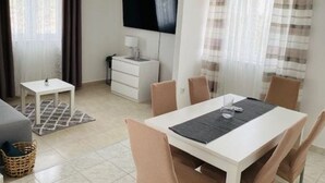 A modern living room with a white sofa, a small white table, a TV mounted on the wall, and a dining area with a white table and chairs. - Villa Brazza (Milna)