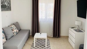 A small, well-lit room with a grey sofa, a white table, and a white dresser. There is a window with brown curtains, and a painting is visible on the wall. - Villa Brazza (Milna)