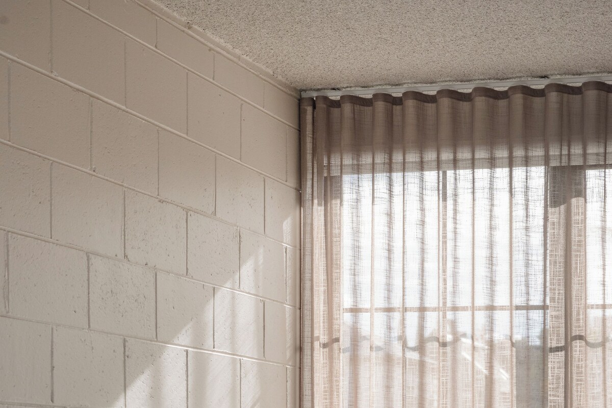 A hotel room with a brick wall, a window with sheer curtains, a round table with a vase of flowers, and a bed with white linens.