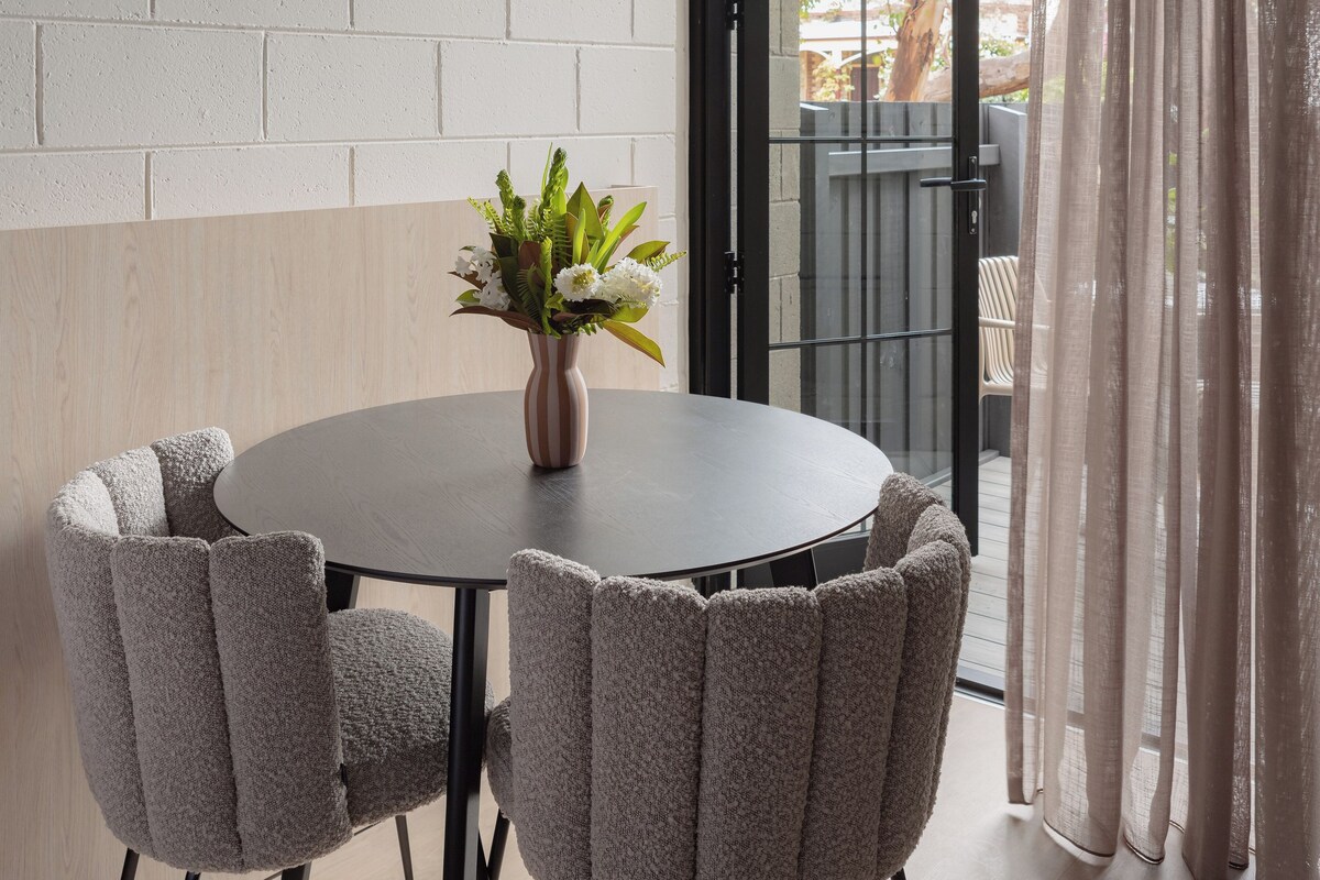 A dining area with a round table, four upholstered chairs, a vase with flowers, and a view through an open door to an outdoor space.