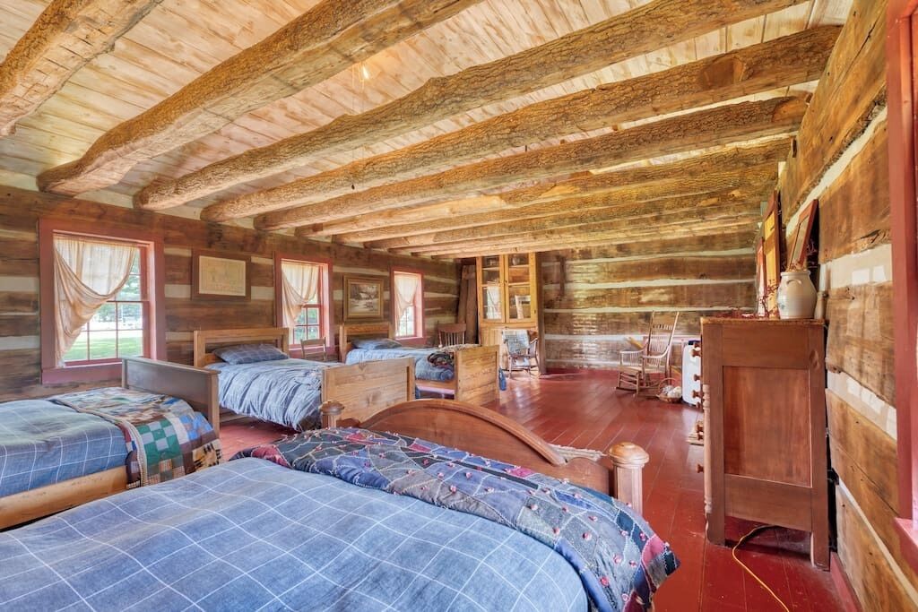 Una cabaña rústica con paredes de madera y techo inclinado. Hay tres camas con colchas, un tocador de madera y una mesita.
