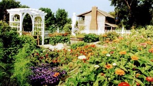 A garden with a white pergola, a small building, and a white picket fence. - Vintage 1850's Log Cabin Glamping (Flora)